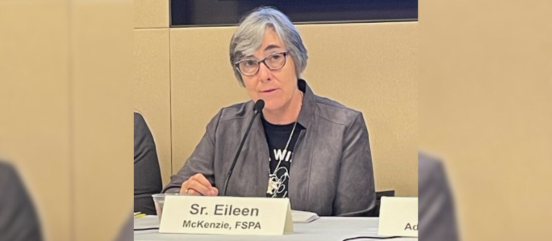 woman in black glasses and shirt with gray hair and jacket sits before a tan background, microphone and Sir. Eileen McKenzie, FSPA