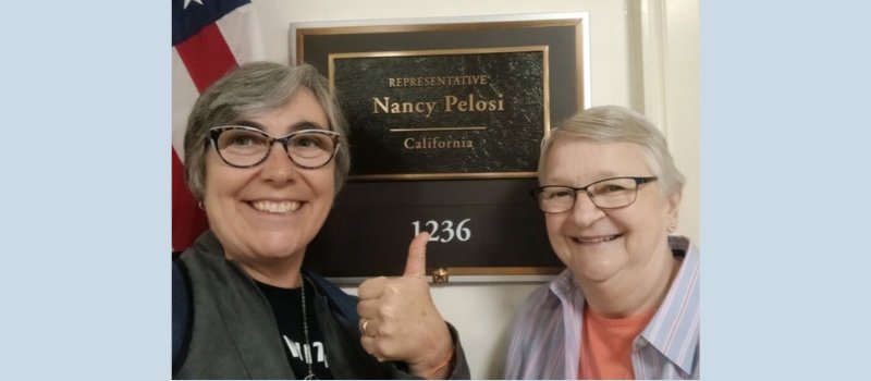 woman with gray hair and sweater, black glasses and t-shirt giving thumbs up and women with gray hair, black glasses, coral t-shirt and blue button up in front of the American flag and plaque for Nancy Pelosi’s office