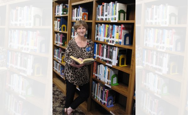 woman with leopard print blouse and short brown hair holds open book in front of shelves of books in library