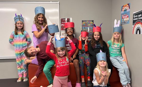 eight little girls and one little boy dressed in colorful clothes with red and blue paper hats in classroom with white board and posters