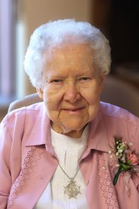 An elderly woman with short white hair looks into the camera, wearing a pink jacket over a white blouse, a silver necklace, and a small pink corsage, seated indoors with a softly blurred background.